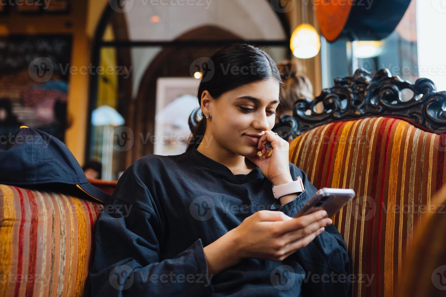 Söt ung flicka vilar på en stor mjuk stol i en Kafé, chattar på de telefon foto