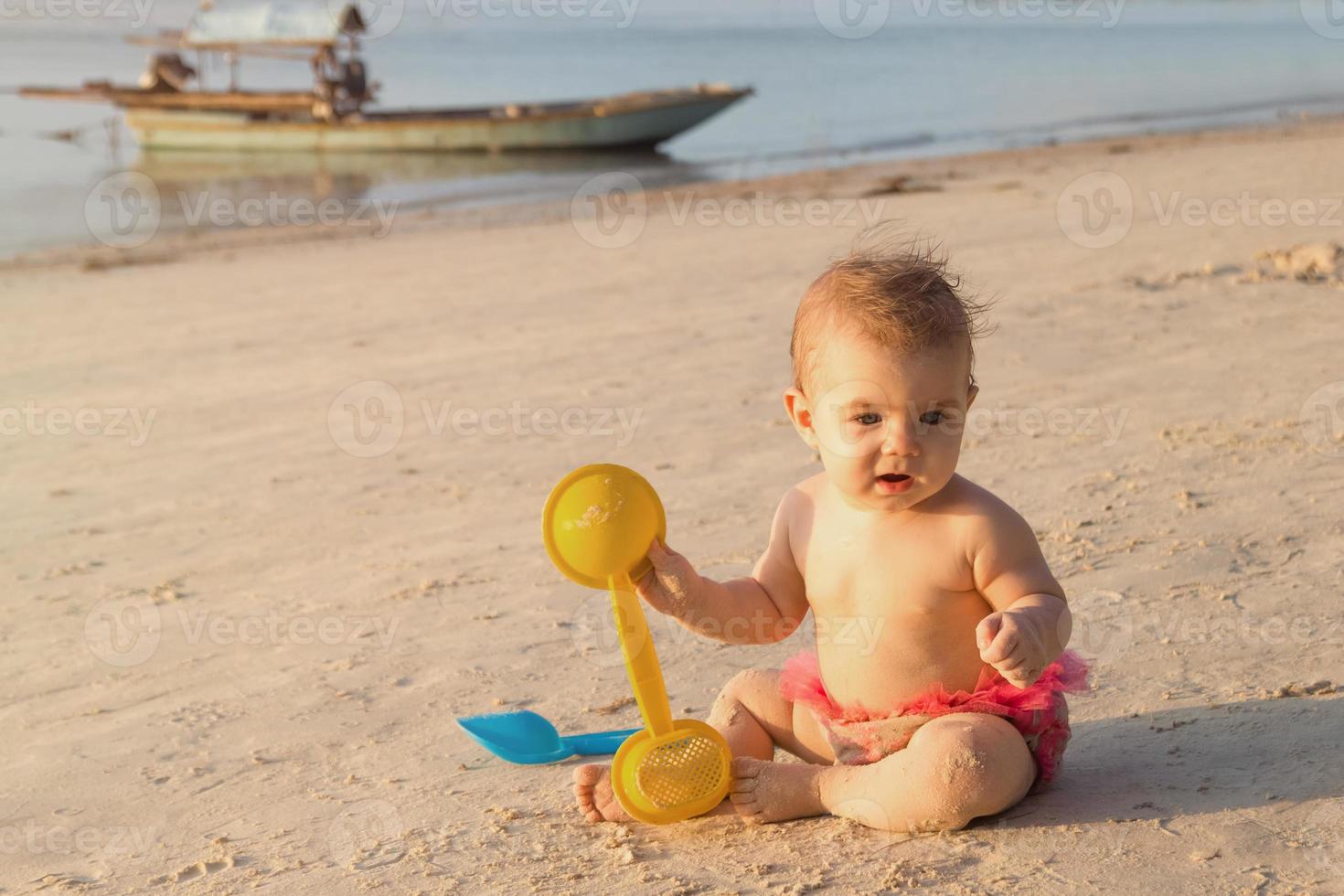 söt liten bebis flicka är Sammanträde på en sandig strand nära till hav i solnedgång solljus och spelar med plast leksaker. foto