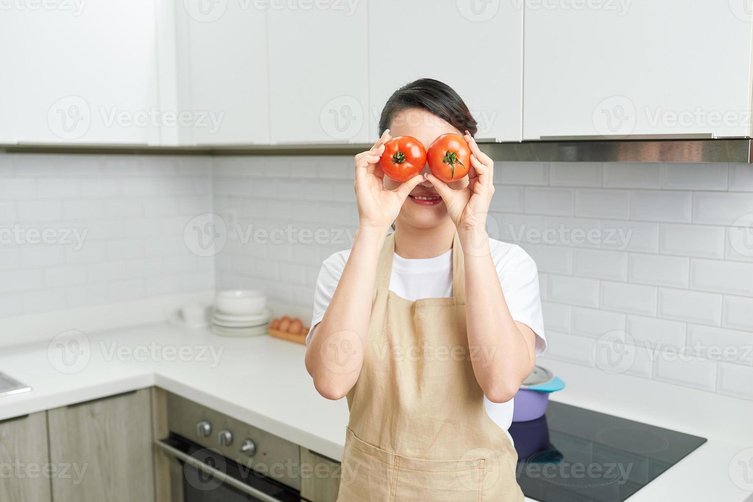 Foto av hemmafru attraktiv lady vapen innehav två stor tomat dölja ögon lekfull humör njut av morgon- matlagning gott middag ha på sig förkläde stå modern kök inomhus