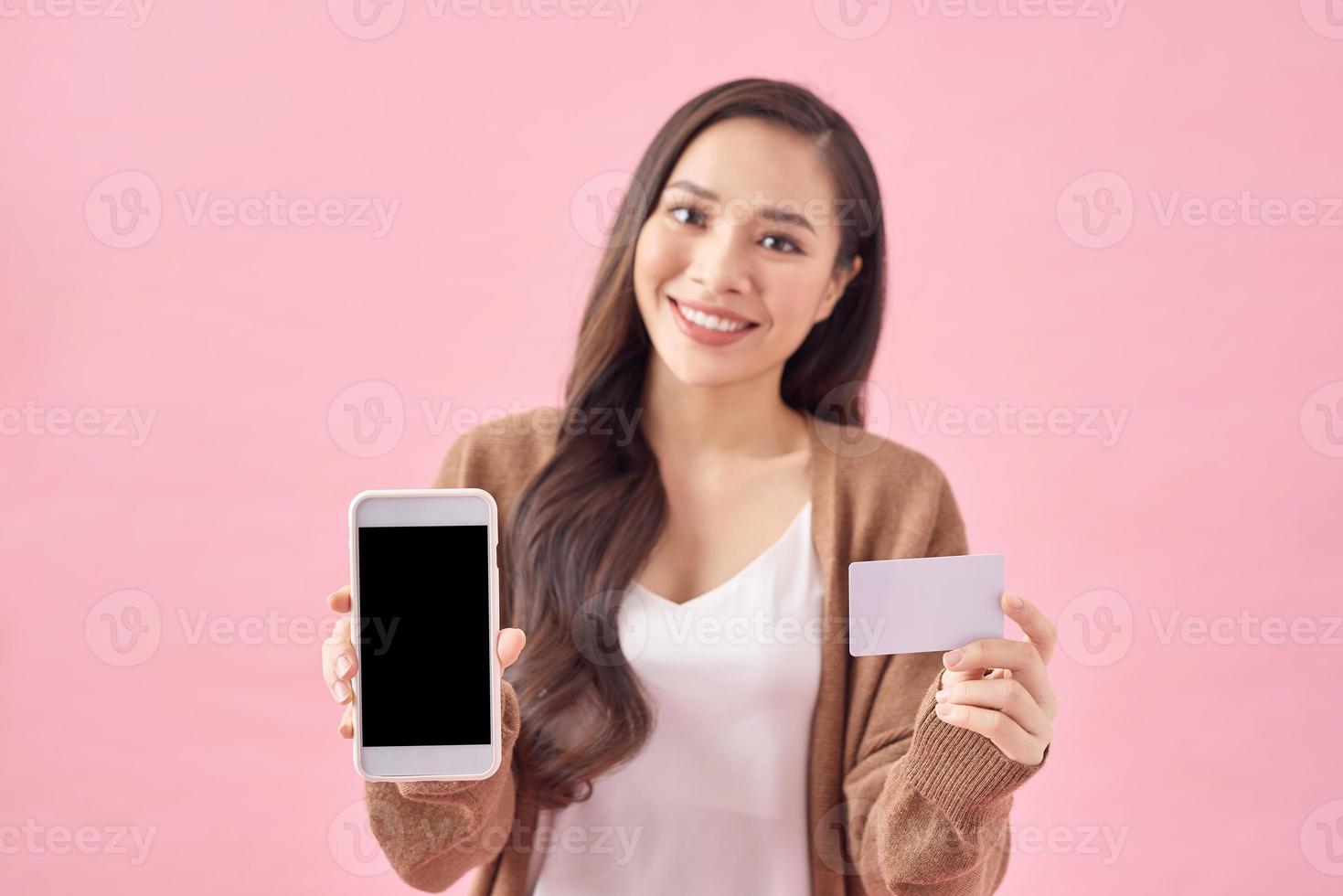 bild av Lycklig ung asiatisk lady stående isolerat över rosa bakgrund. ser på kamera medan använder sig av telefon och innehav debitera kort. foto