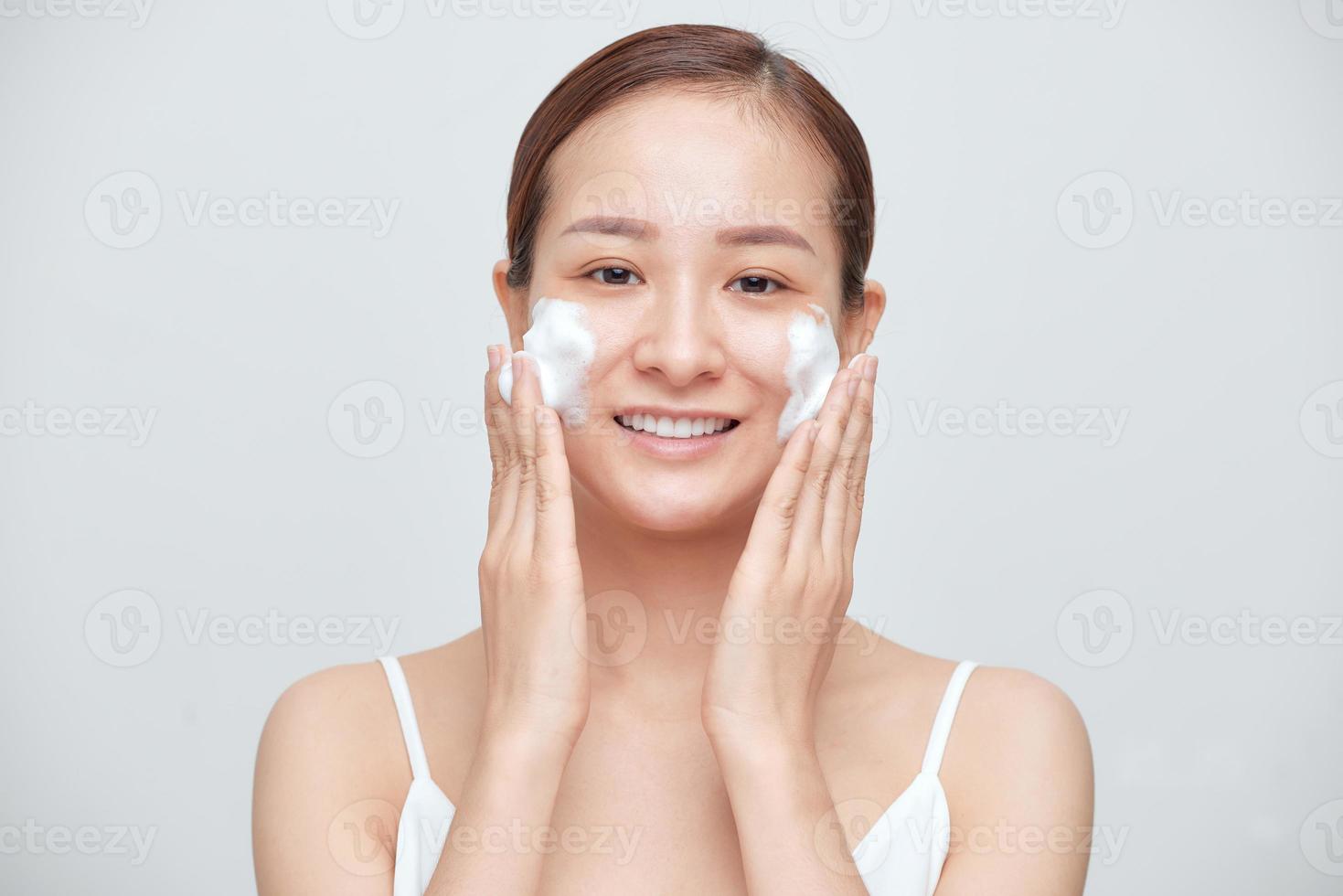 Lycklig asiatisk kvinna tvätta henne ansikte med tvål skum över vit bakgrund. foto