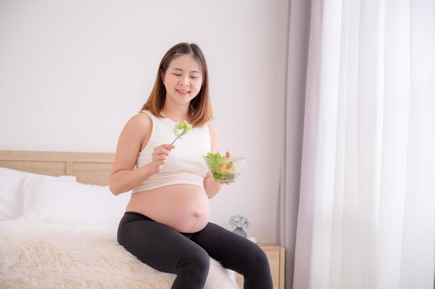 ett asiatisk kvinna sitter i henne sovrum äter friska, organisk sallader medan hon är gravid fram tills de tid av henne bebis födelse foto
