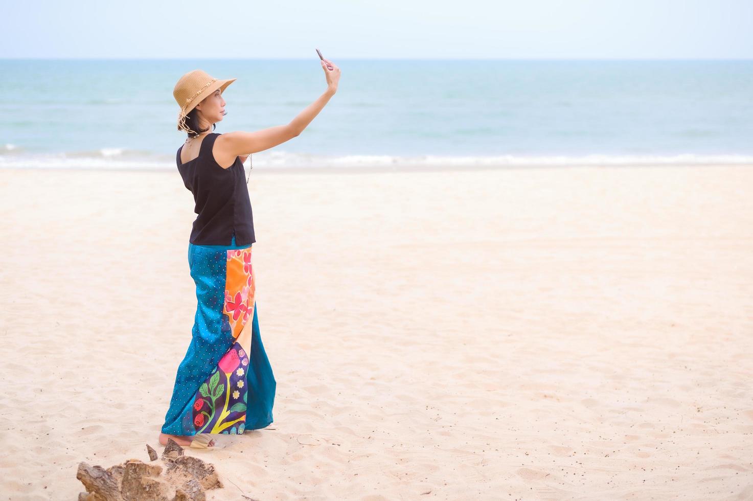 ett asiatisk kvinna står till ta en selfie med henne mobil telefon i de sand under en sommar semester på de strand foto