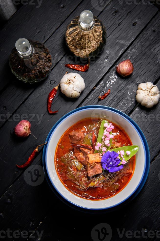 fläsk röd curry eras och dekorerad med grönsaker, örter och kryddor på rustik bakgrund - thai mat foto