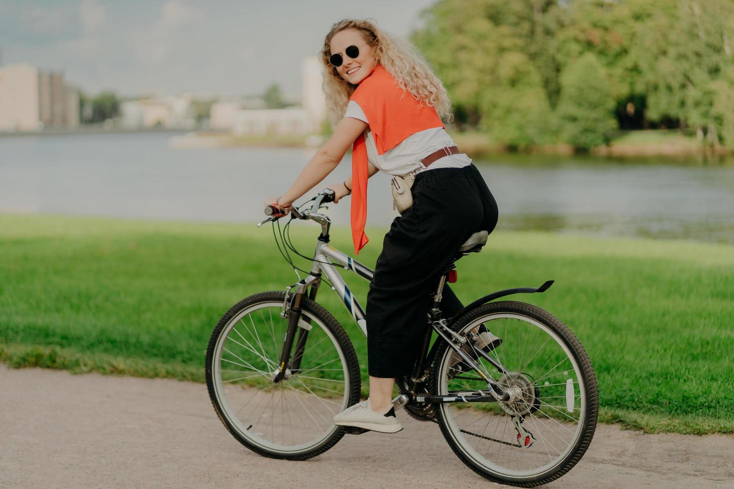 glad ung kvinna njuter av en ny väg på cykel, cyklar bland sjöar, gröna gräsmattor och byggnader långt borta, bär sommarnyanser, avslappnad outfit, vita sneakers, är i bra fysisk form foto