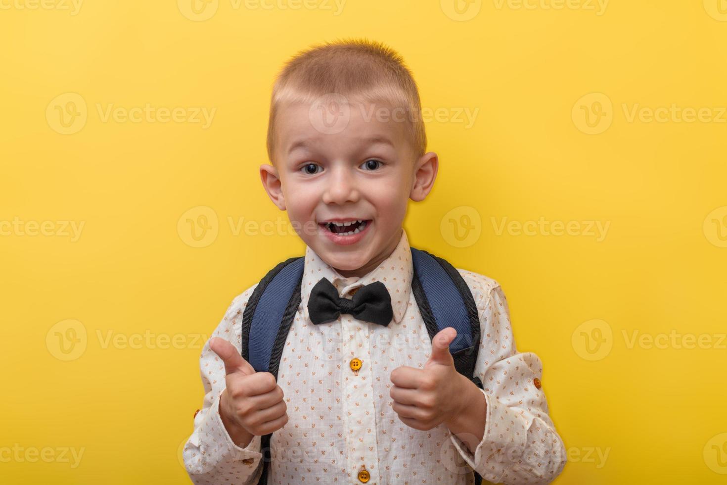 tillbaka till skola. en blond rolig pojke med en ryggsäck i en ljus skjorta på en gul bakgrund visar super, klass och ler. utseende på de kamera. kopia Plats. utbildning. foto