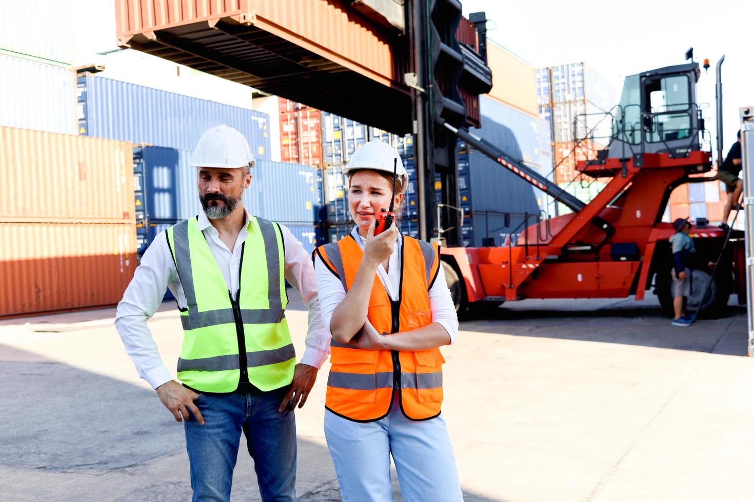 porträtt av två arbetare bär skyddsväst och hjälm diskuterar på logistisk frakt last container varv, senior ingenjör man pratar med vacker ung kvinna kollega på arbetsplatsen. foto