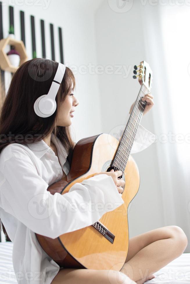 porträtt ungt leende glad vacker asiatisk kvinna koppla av sovrum. kvinna med hörlurar spelar akustisk gitarr i det vita sovrummet foto