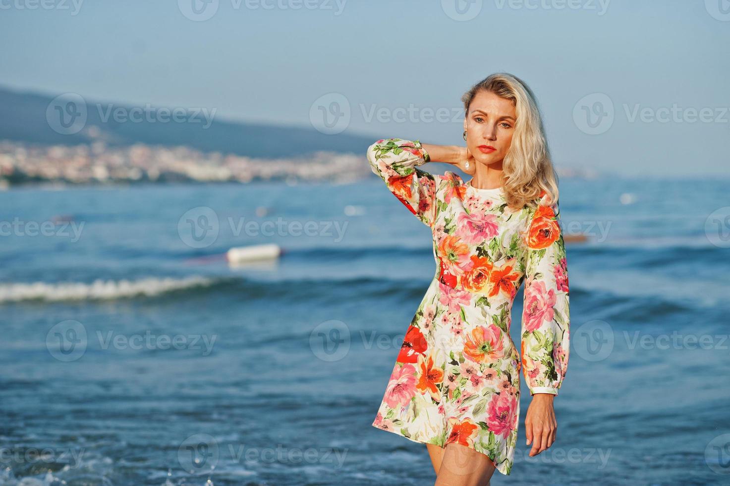 blond kvinna som bär klänning står på sandstranden vid havet och njuter av utsikten över solnedgången. foto