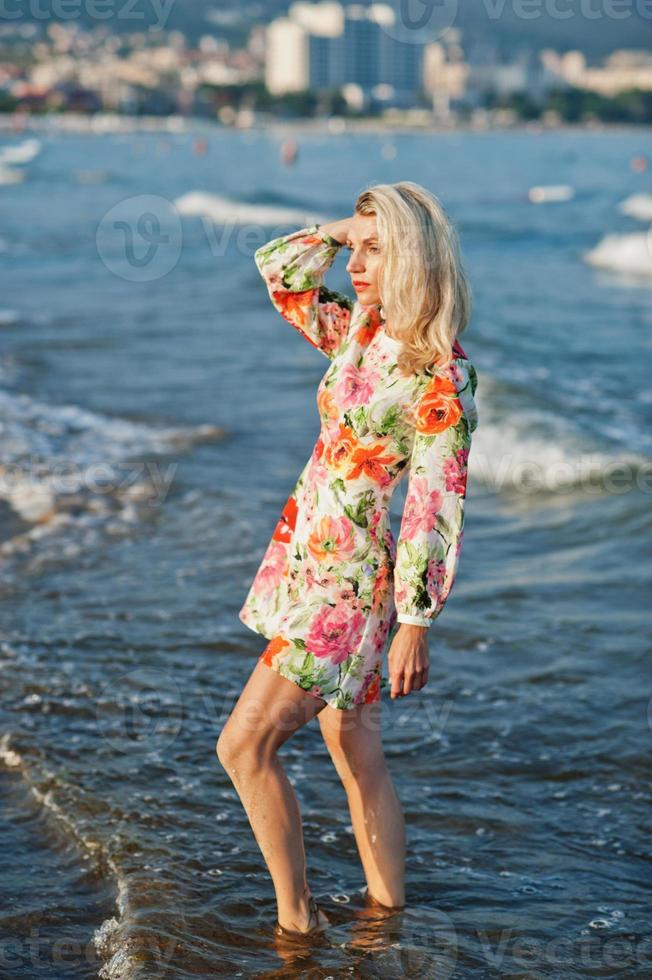 blond kvinna som bär klänning står på sandstranden vid havet och njuter av utsikten över solnedgången. foto