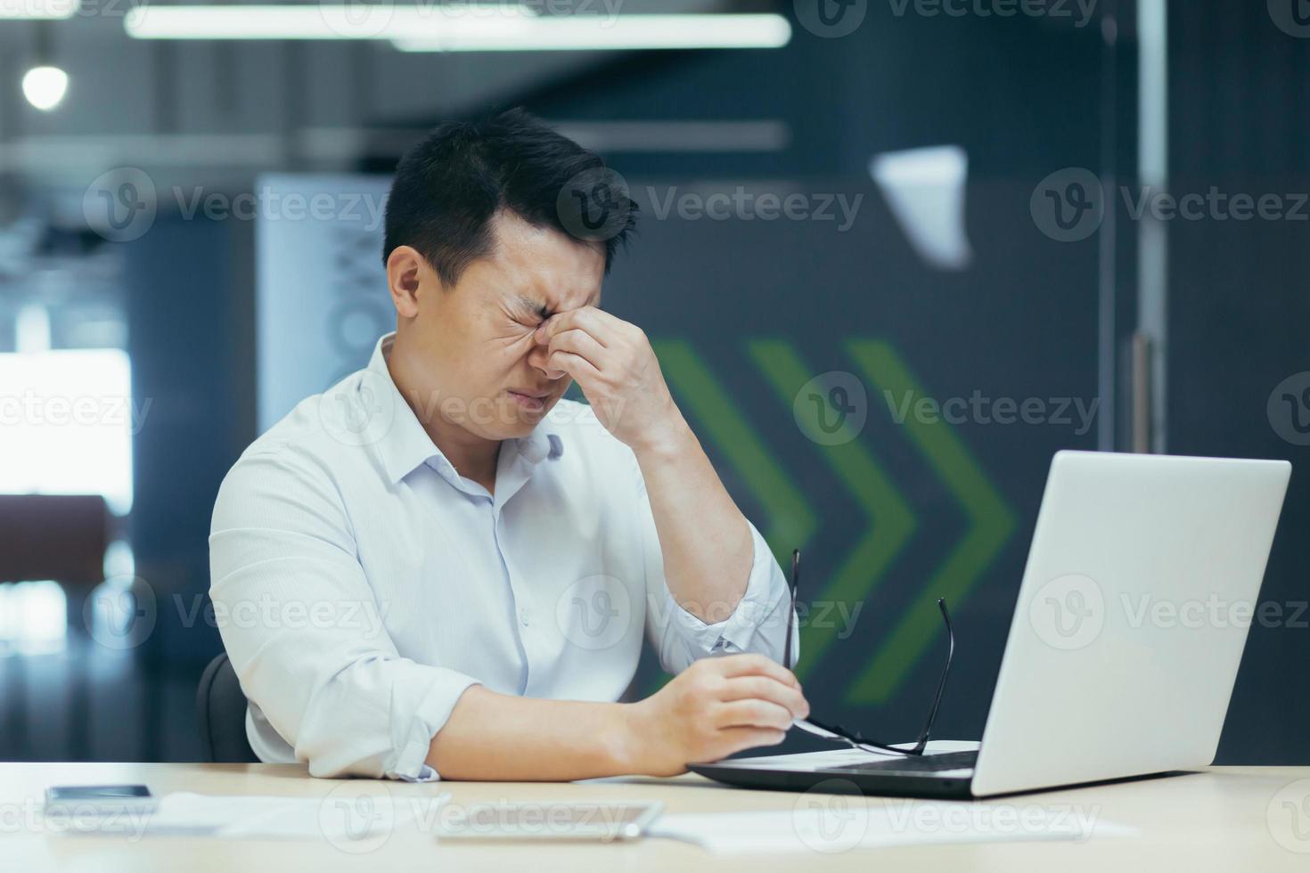 trött asiatisk affärsman arbetssätt sent i kontor, öm ögon, man med bärbar dator. foto