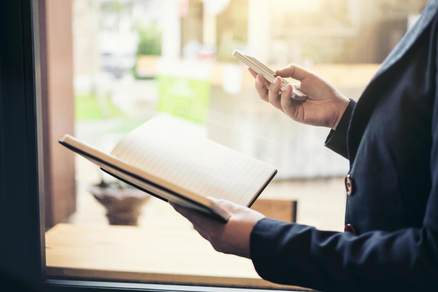 ung attraktiv affärskvinna som står nära fönstret, arbetar på smartphonen med rapportbok, ledarflicka som arbetar, lärande online foto