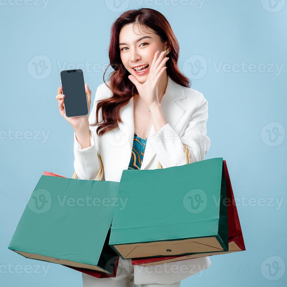 halv kropp av skön asiatisk företag kvinna bär vit kostym öppen mun höjning händer skrikande meddelande som visar mobil telefon Ansökan och handla väska på isolerat blå bakgrund. foto