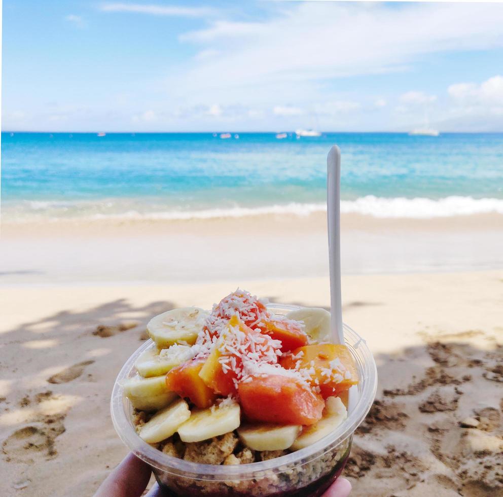 person som håller en gul och orange fruktsallad i en kopp på en strand foto