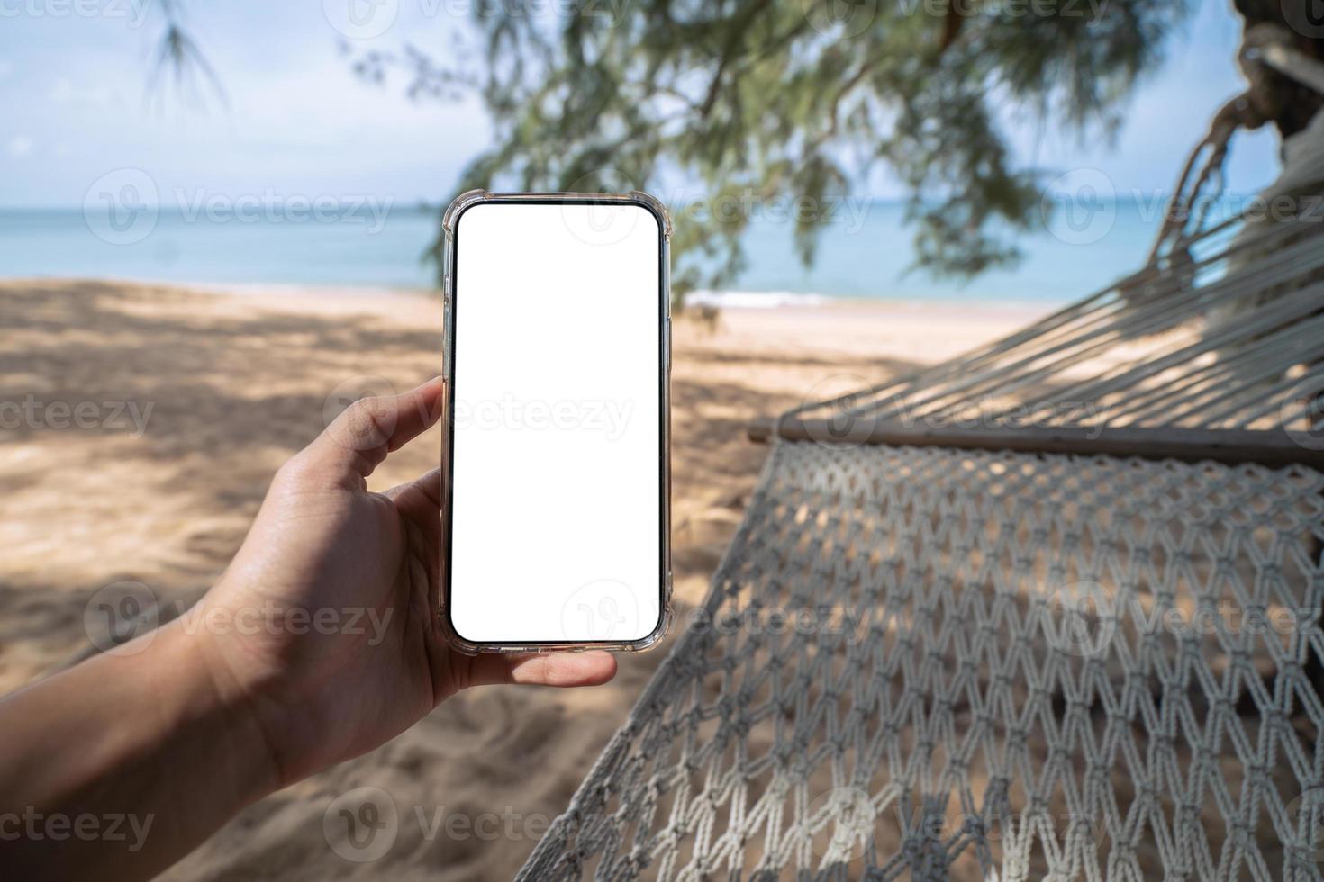 hand innehav falsk upp mobil med vit sceen medan om på hängmatta gunga mellan träd på de strand. foto