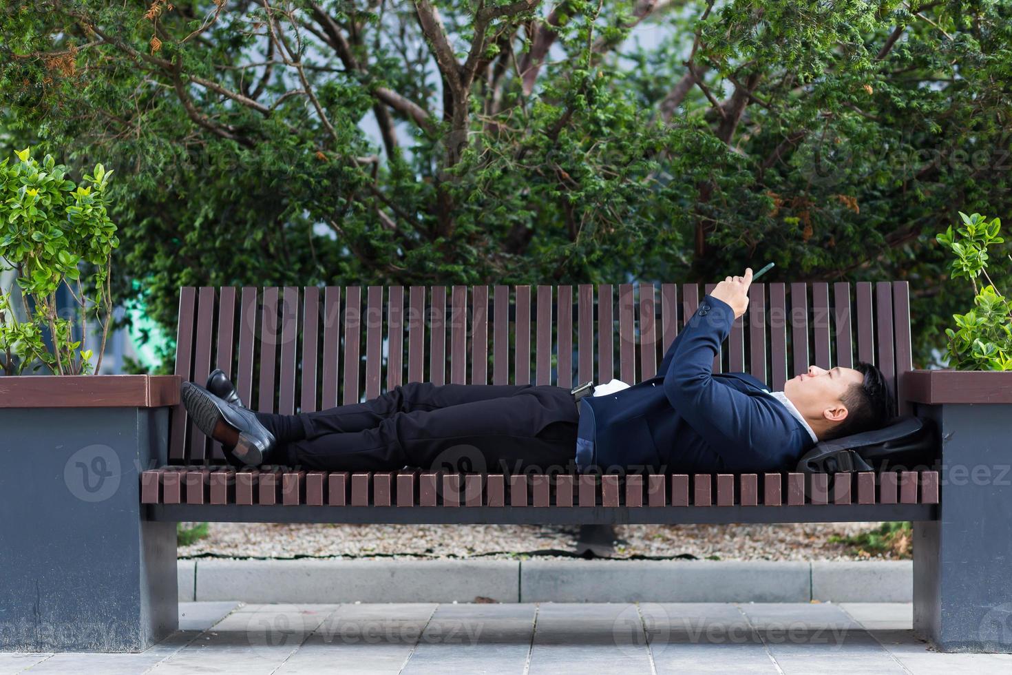 manlig asiatisk affärsman liggande på en bänk och vilar efter arbete användningar en mobil telefon foto