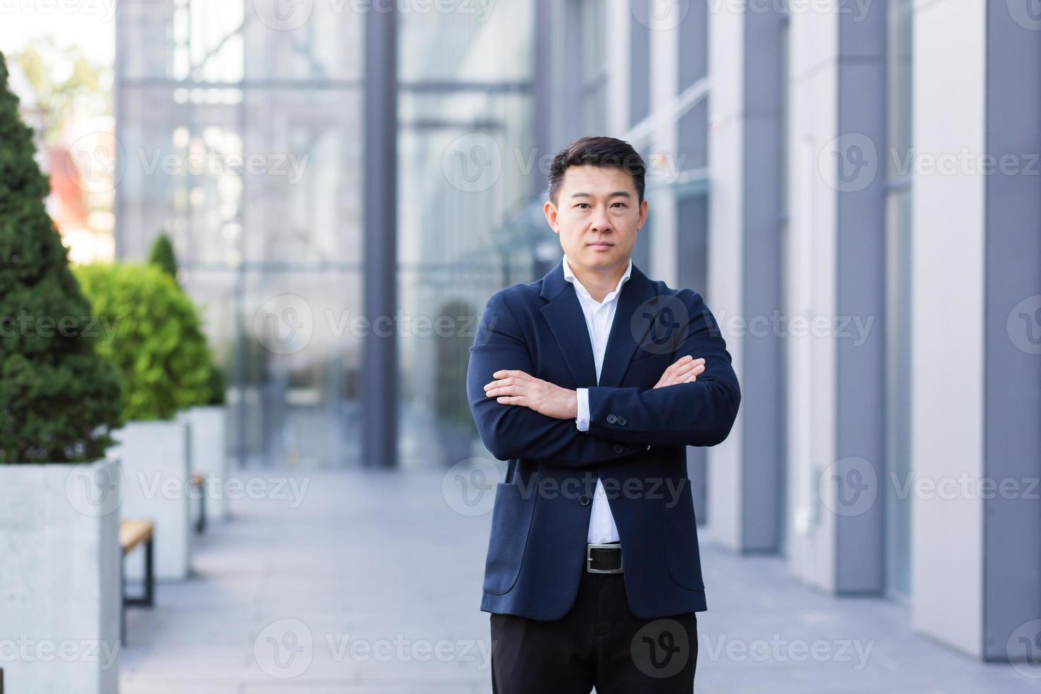 framgångsrik asiatisk chef nära kontor ser på kamera med korsade vapen av allvarlig affärsman foto