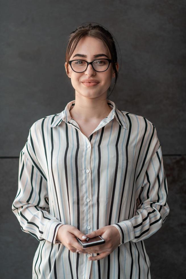 börja affärskvinna i skjorta med en glasögon använder sig av smartphone medan stående i främre av grå vägg under ha sönder från arbete utanför foto