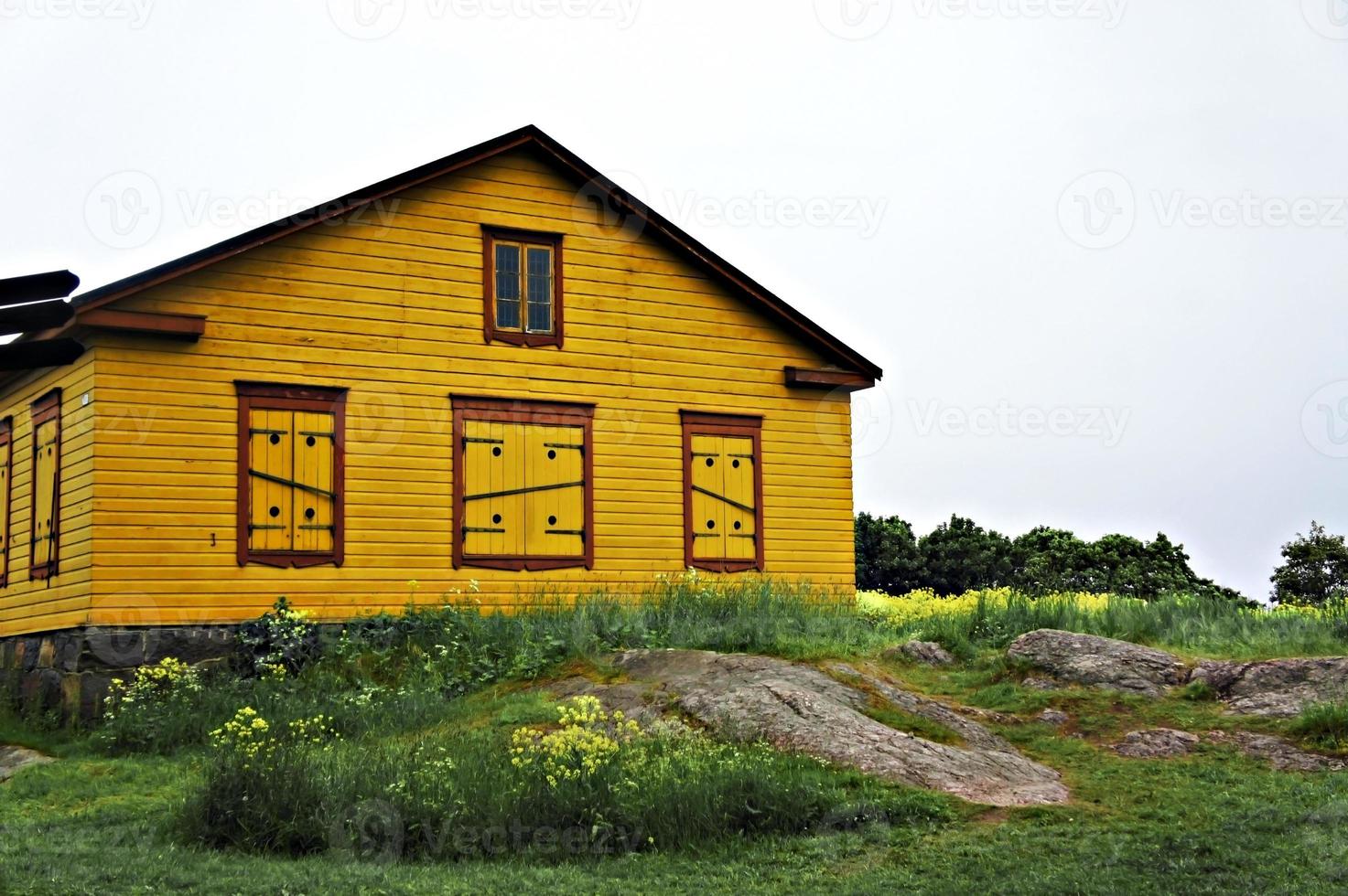 gul trä- hus med stängd jalusier bland grön gräs, gul blommor och stor stenar foto