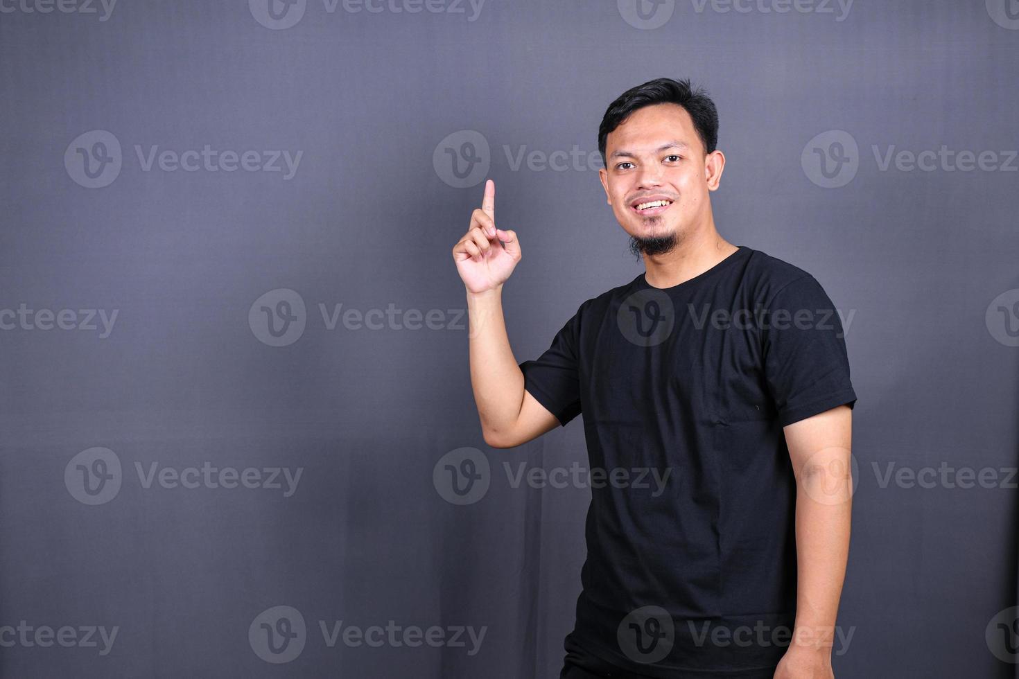 porträtt av en Lycklig ung asiatisk man i svart t-shirt pekande fingrar bort på kopia Plats isolerat över grå bakgrund foto