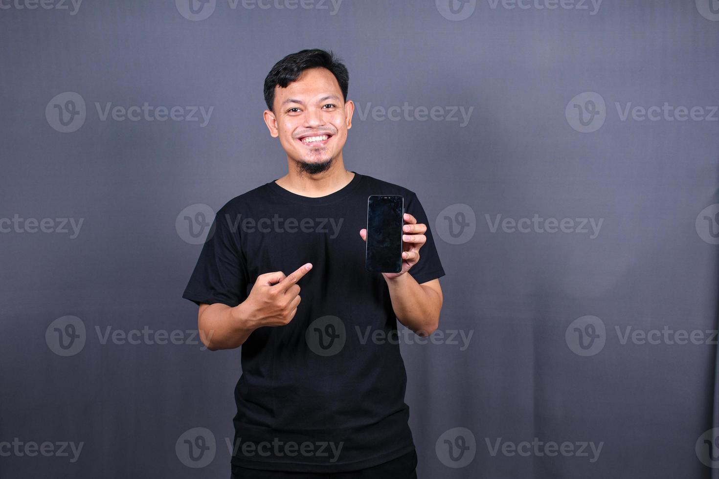 porträtt av ung asiatisk man bär svart t-shirt stående isolerat över grå bakgrund, pekande finger på mobil telefon foto