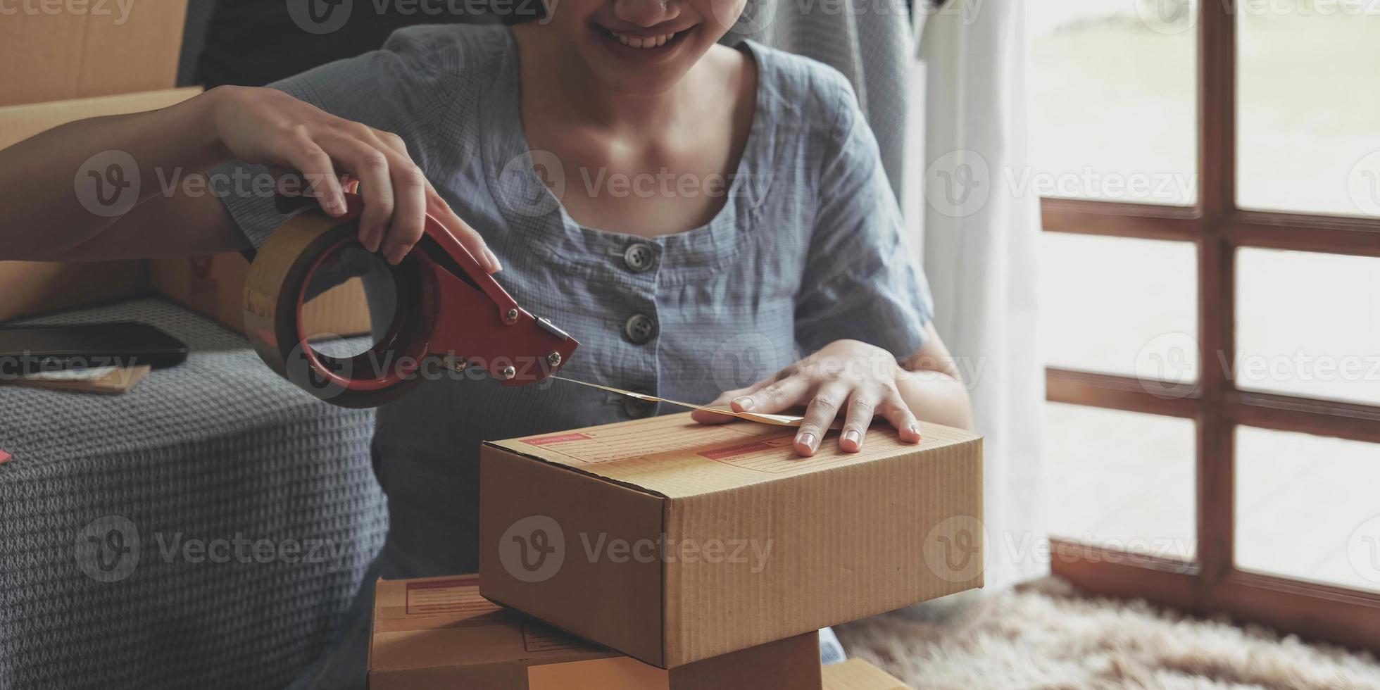 små företag entreprenör sme, asiatisk ung kvinna, tjej ägare förpackning produkt, kontroll paket för leverans till kund, använder sig av scotch tejp till täta de låda, arbetssätt på Hem. handlare uppkopplad e handel foto