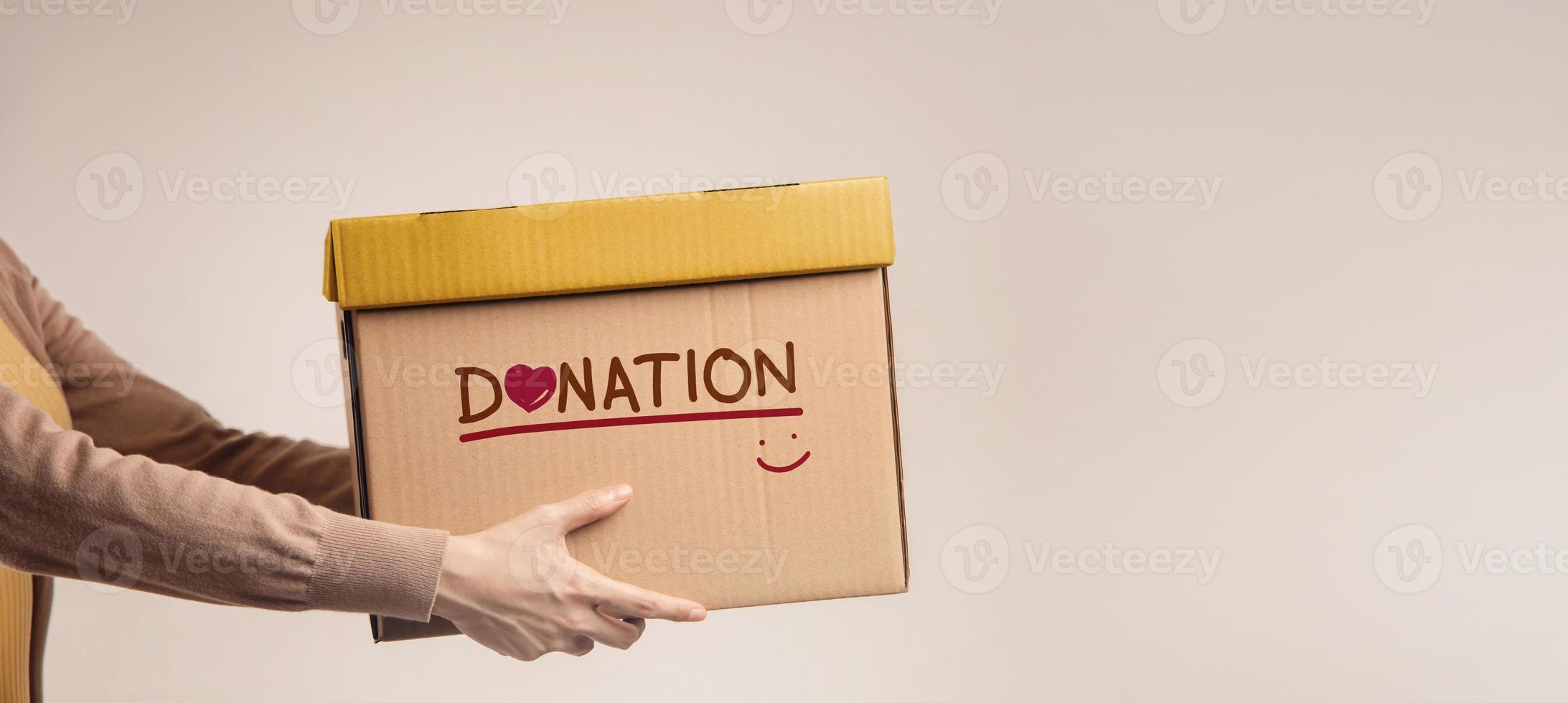 donation begrepp. kvinna med låda av saker för donera med donation märka, leende och hjärta. stående mot de walll. sida se foto