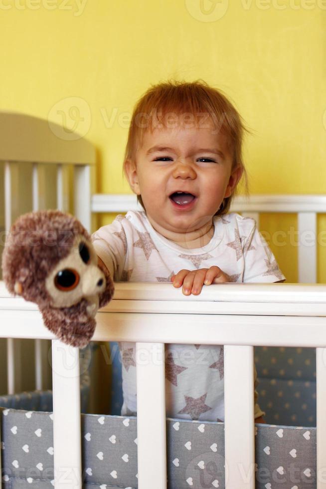 Lycklig ett år gammal flicka är spelar med plysch leksak efter vakna upp i de morgon- i henne säng. foto