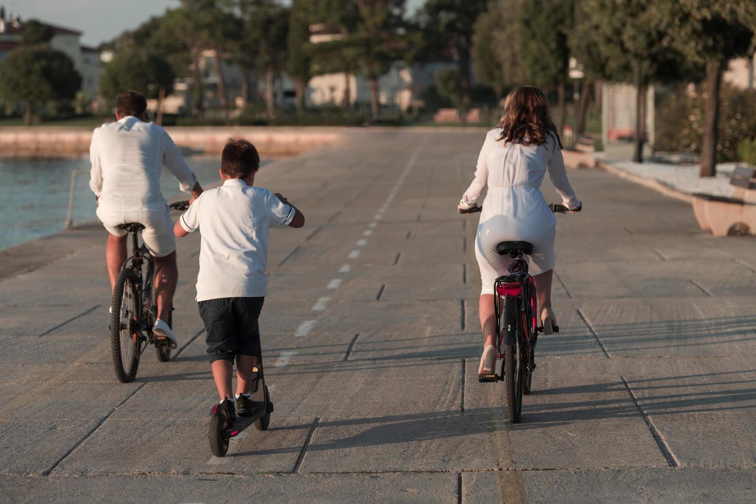 Lycklig familj njuter en skön morgon- förbi de hav tillsammans, föräldrar ridning en cykel och deras son ridning ett elektrisk skoter. selektiv fokus foto