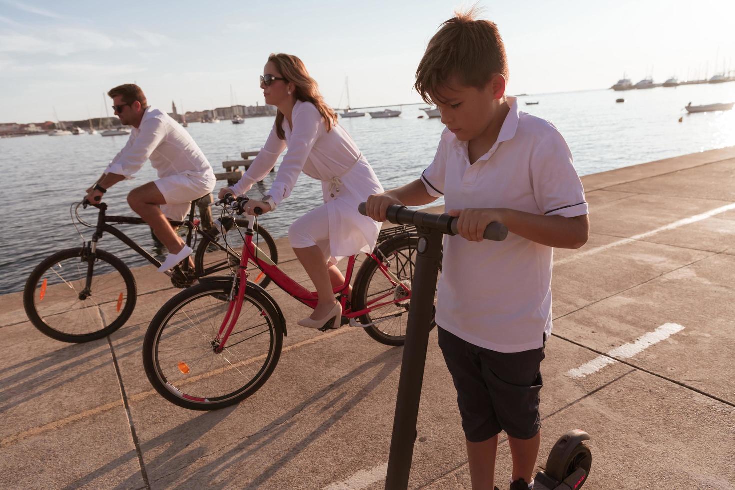 Lycklig familj njuter en skön morgon- förbi de hav tillsammans, föräldrar ridning en cykel och deras son ridning ett elektrisk skoter. selektiv fokus foto