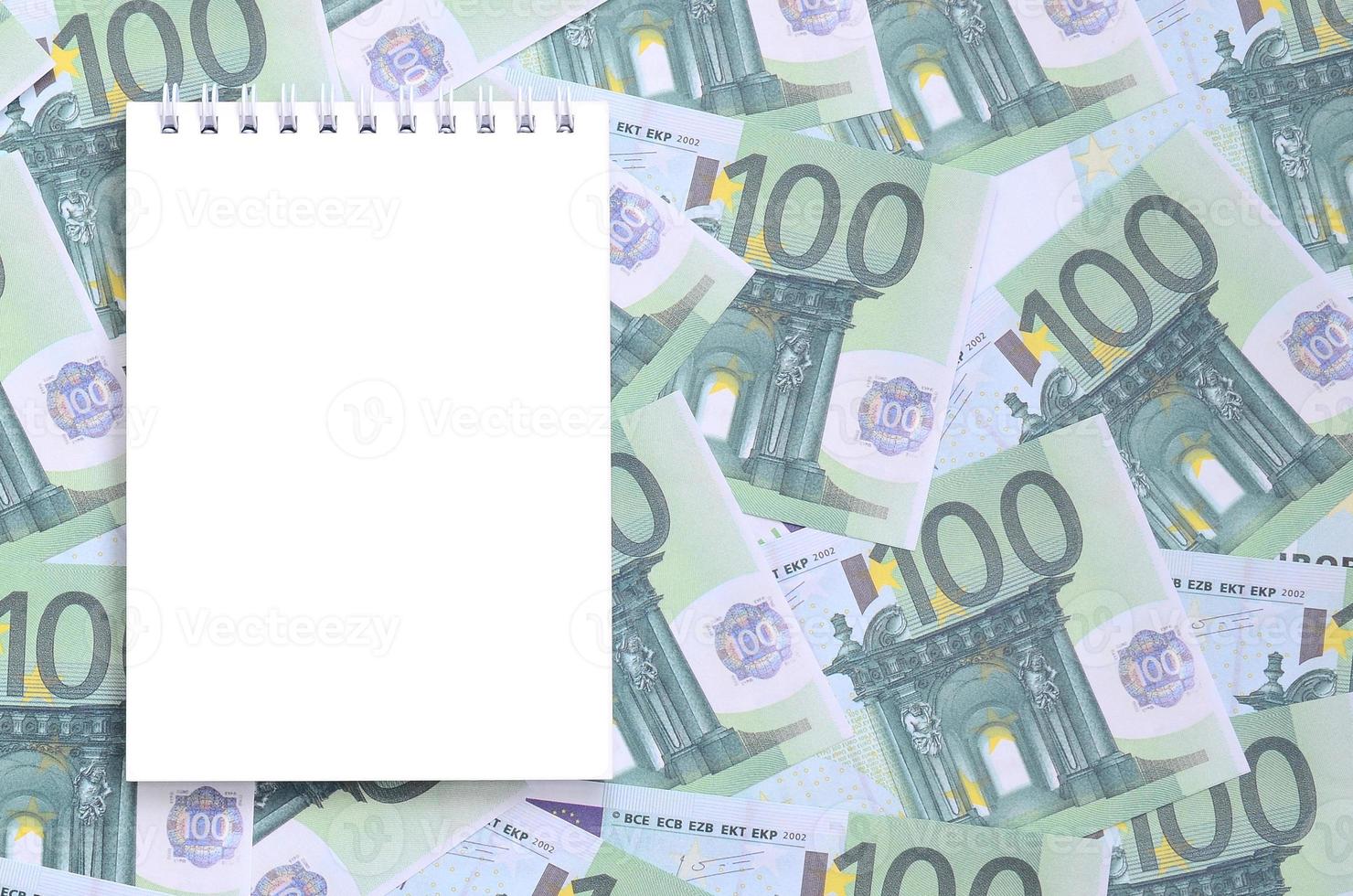 vit anteckningsbok med rena sidor liggande på en uppsättning av grön monetär valörer av 100 euro. en massa av pengar former ett oändlig högen foto