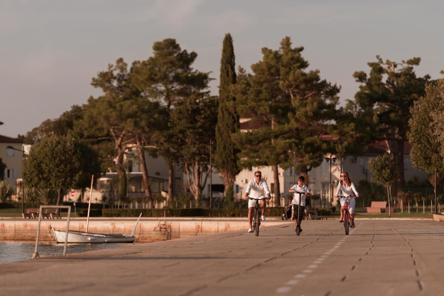 Lycklig familj njuter en skön morgon- förbi de hav tillsammans, föräldrar ridning en cykel och deras son ridning ett elektrisk skoter. selektiv fokus foto