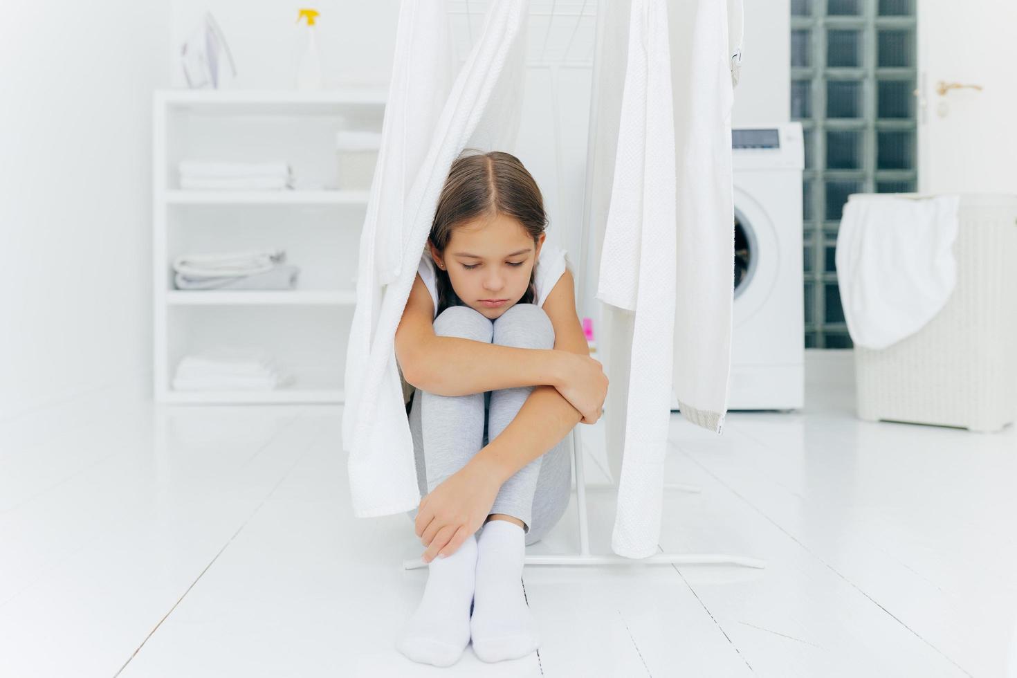 ensam omtänksam ledsen liten flicka sitter på golvet i tvättrummet nära torktumlare, bär byxor och vita strumpor, tvättmaskin, tvättkorg och konsol i bakgrunden, väntar på föräldrar foto