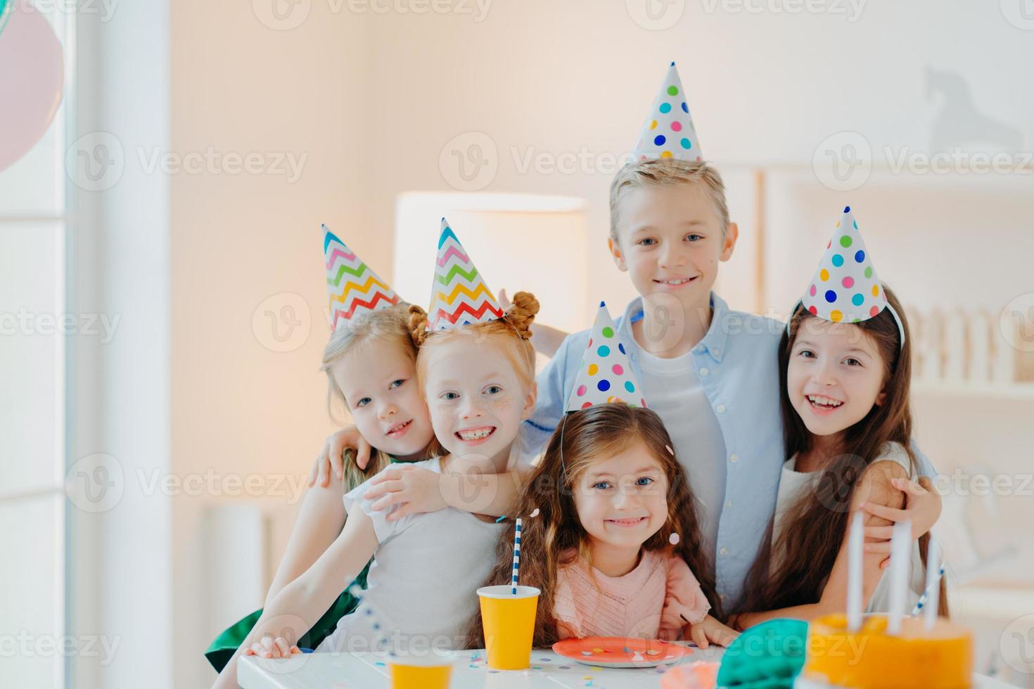 en grupp bedårande barn bär festhattar, omfamnar och har roligt, firar födelsedag, poserar i ett dekorerat rum, samlas nära festbordet, ha roligt och myser och tittar glatt på kameran foto