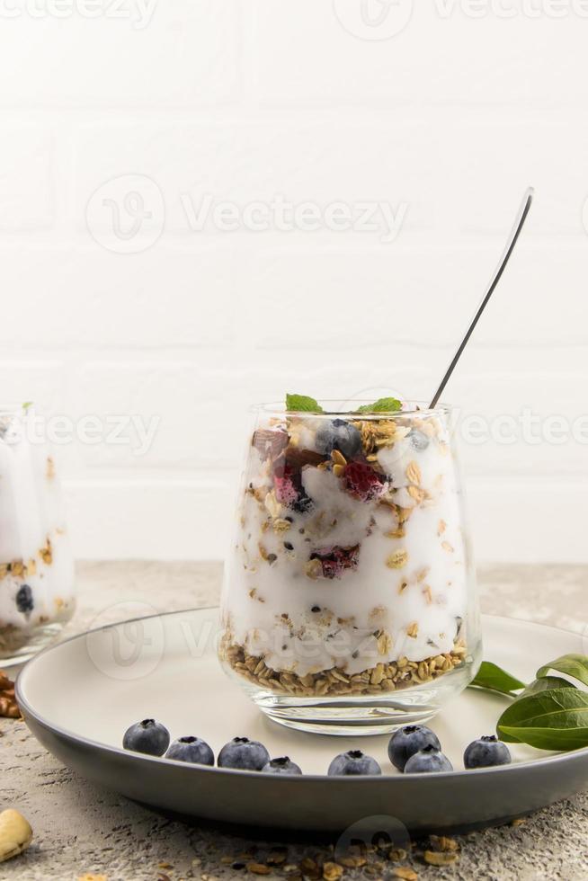 en glas av ljus yoghurt, mysli, färsk bär, nötter står på en keramisk tallrik med blåbär och grön löv. diet frukost. foto