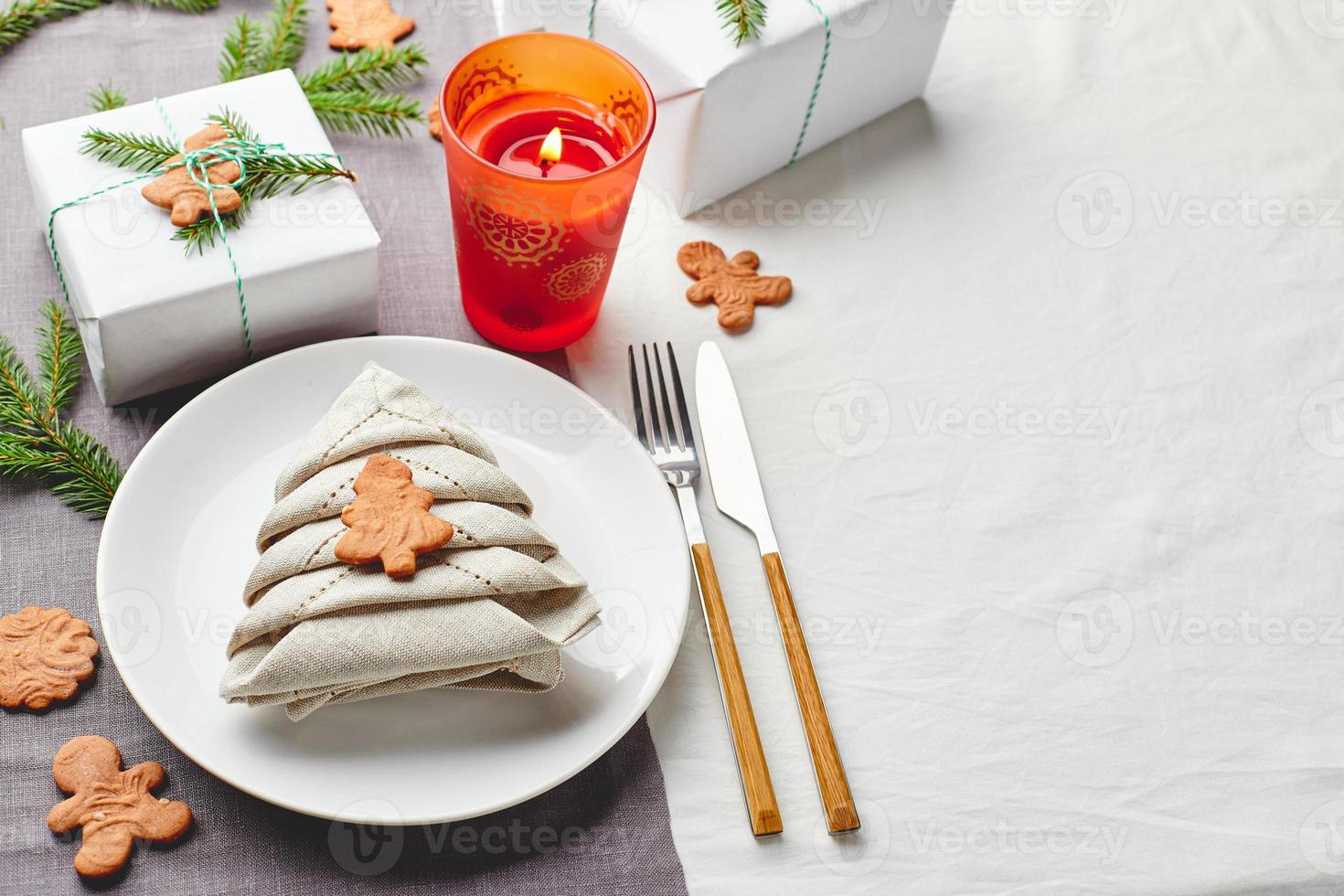 servett i de form av en jul träd på en tallrik på vit bordsduk med gåvor och dekorationer med gran kvistar och pepparkaka småkakor foto