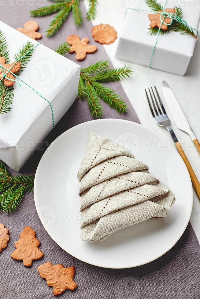 servett i de form av en jul träd på en tallrik på vit bordsduk med gåvor och dekorationer med gran kvistar och pepparkaka småkakor foto