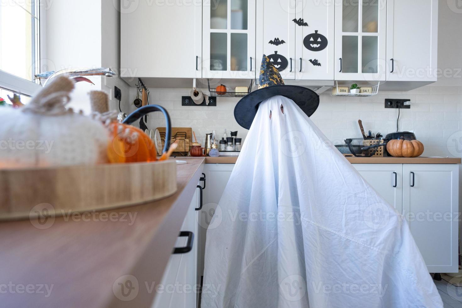 en barn i ark med Skära ut för ögon tycka om en spöke kostym i de kök dekorerad för de halloween Semester. en snäll liten rolig spöke. halloween fest foto