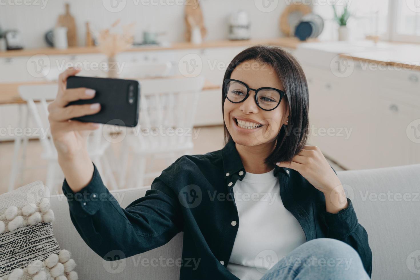 leende flicka bloggare innehav smartphone, samtal förbi video ringa upp, tar selfie foton, Sammanträde på soffa foto