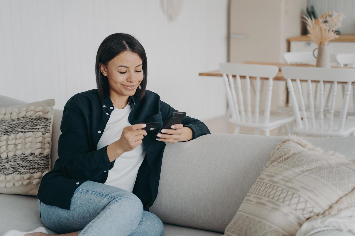 leende kvinna innehav Bank kreditera kort, telefon gör framgångsrik kontantlös betalning på soffa på Hem foto