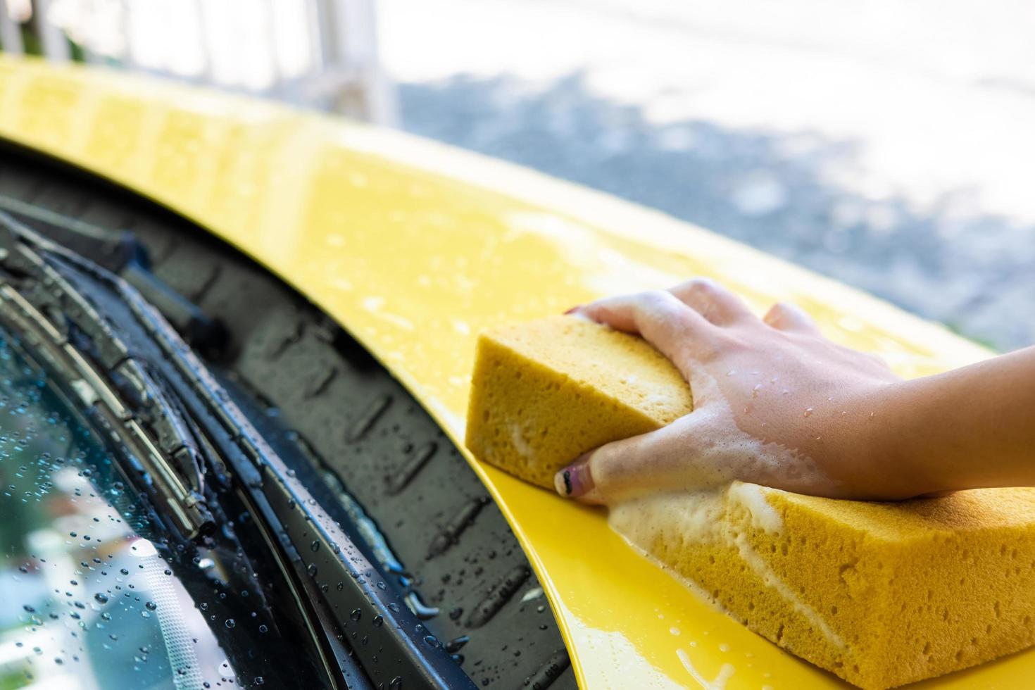 selektiv fokus på kvinnor hand använder sig av svamp till tvätta och rena färgrik gul bil med tvål bubbla på Hem. själv bil vård underhåll aktivitet. foto