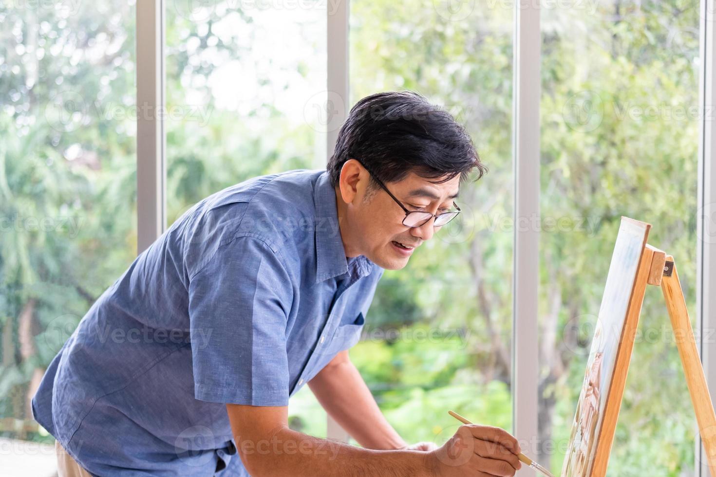 inomhus- skott av senior asiatisk män använder sig av borsta och olja Färg till måla på målning duk. med leende på hans ansikte. Lycklig pensionering begrepp för aktiva senior. med suddig grön natur bakgrund. foto