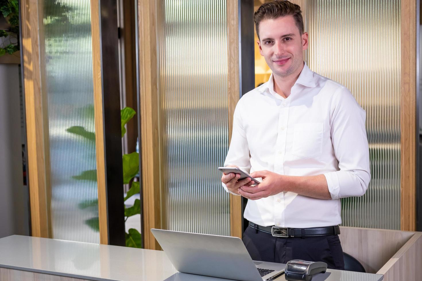 ung caucasian män klänning i formell vit skjorta stående på de reception disken med kreditera kort läsare maskin. medan innehav smartphone enhet med leende på hans ansikte. inköp punkt. foto