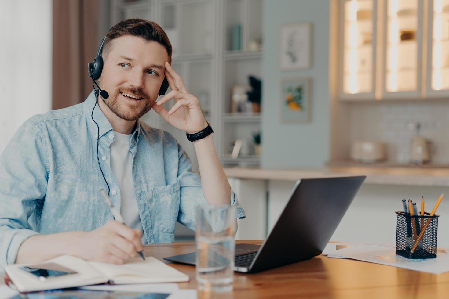 fundersam eftertänksam man gör anteckningar använder bärbar dator headset skriver ner anteckningar i anteckningsblock tillbehör kundsupport löser klientproblem sitter vid skrivbordet i coworking space. hotline chef. foto