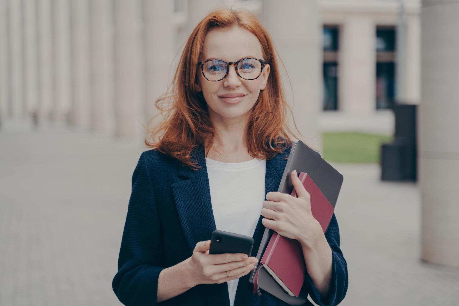 självsäker vacker rödhårig kvinnlig affärskonsult som håller modern smartphone och bärbar dator foto