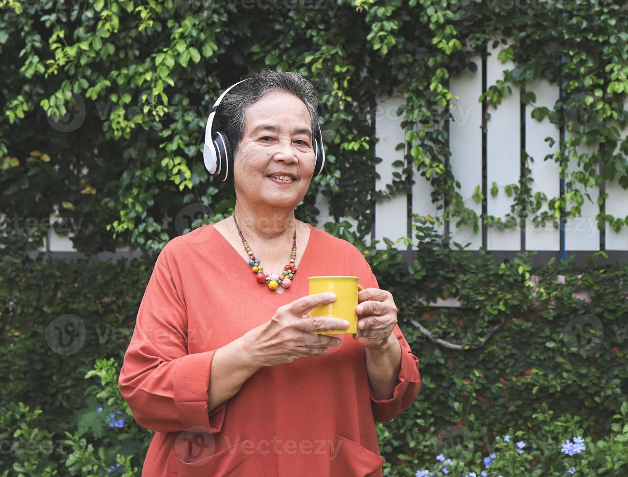Lycklig och friska äldre asiatisk kvinna innehav gul kopp av kaffe stående i de trädgård, lyssnande till de musik från hörlurar, leende och ser på kamera. foto
