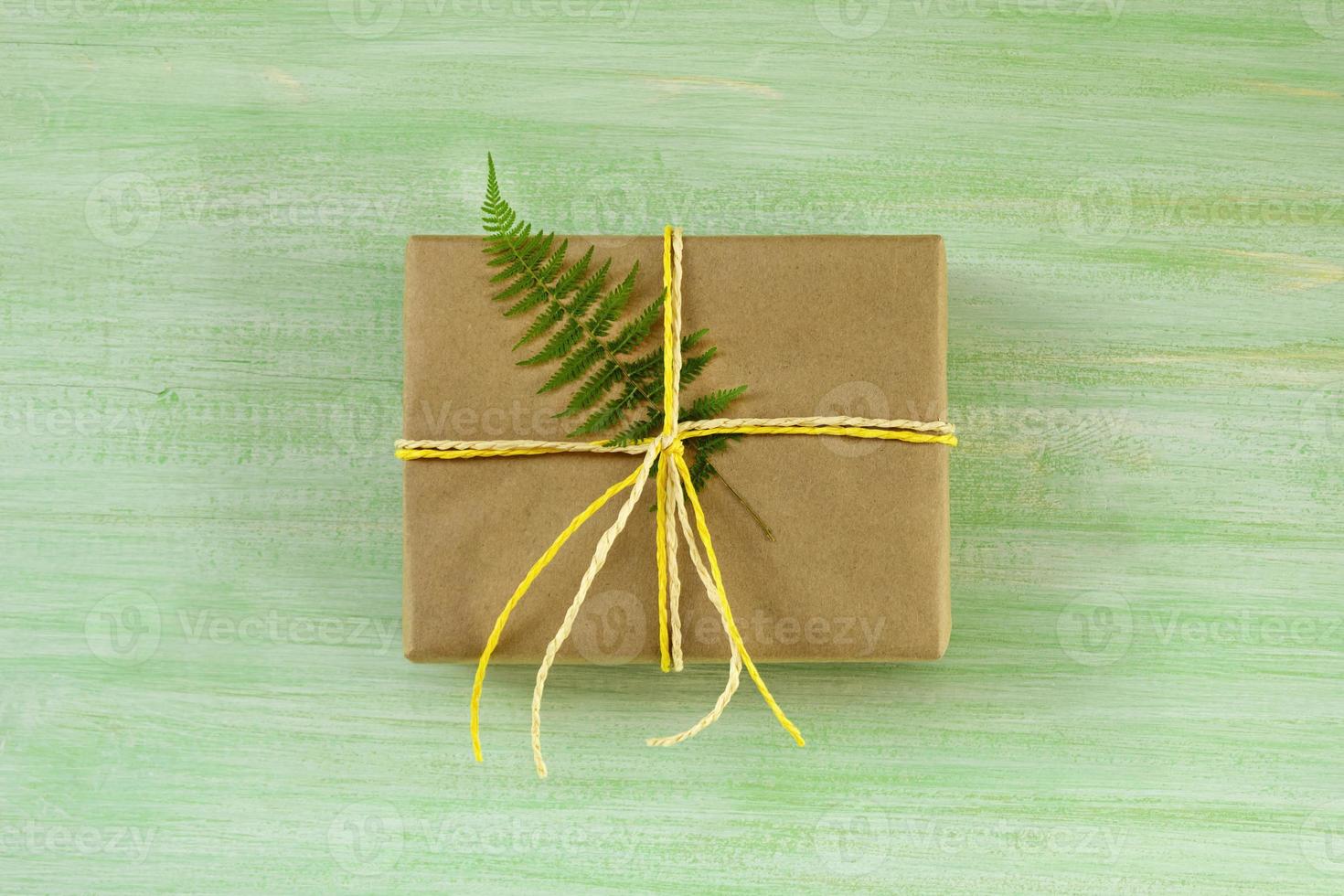 gåva låda insvept av hantverk papper och vit och gul band med ormbunke blad på en grön trä- bakgrund, topp se. foto