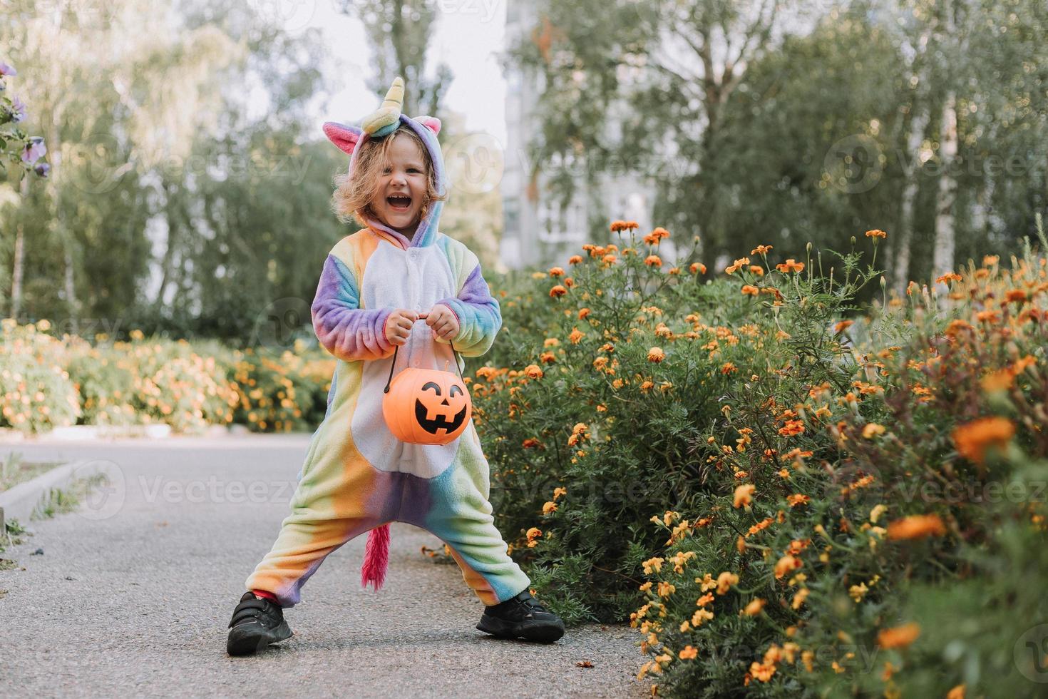 söt liten flicka i en regnbåge enhörning kostym för halloween går till samla sötsaker i en pumpa korg i en bostads- område. barn promenader i de utomhus. lura eller behandla. livsstil. kigurumi foto