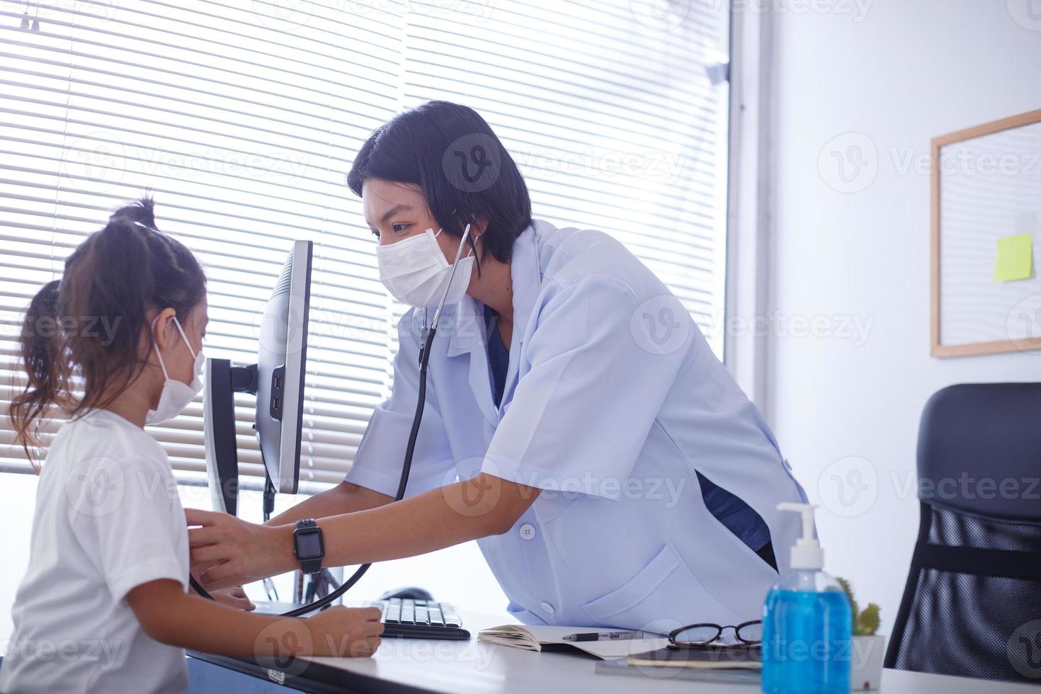 ett asiatisk kvinna läkare bär en kirurgisk mask är kontroll de barn patientens hjärta Betygsätta med en stetoskop. foto