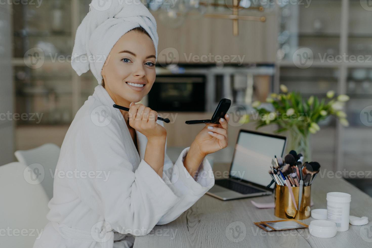 öm härlig kvinna med frisk hud håller en liten spegel och en kosmetisk borste applicerar puder i ansiktet ler tandigt bär mjuk vit morgonrock och handduk poserar mot heminredning. skönhet koncept foto